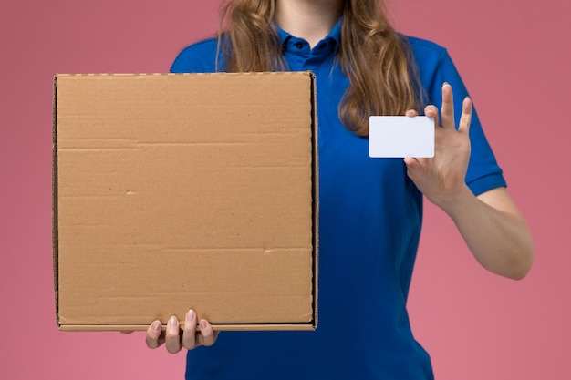 Free photo front close view female courier in blue uniform holding food delivery box and white card on the pink desk service uniform company worker