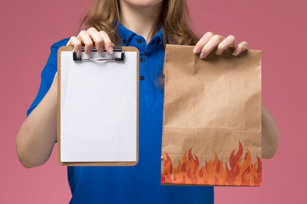 Front close view female courier in blue uniform holding notepad and food package on light-pink desk job worker service uniform company