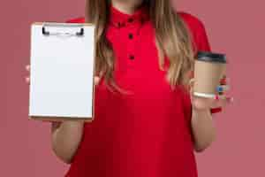 Foto gratuita corriere femminile di vista ravvicinata anteriore in uniforme rossa e tazza di caffè di consegna della tenuta del capo con il blocco note sull'uniforme di consegna del lavoro di servizio del fondo rosa chiaro