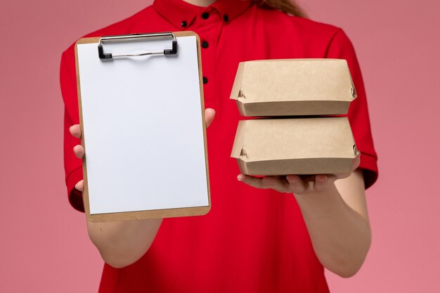 Front close view female courier in red uniform and cape holding notepad and little delivery food packages on light-pink wall, service uniform delivery