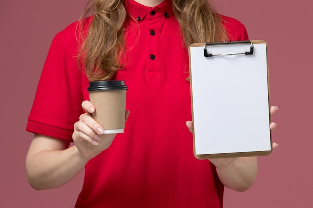 Corriere femminile di vista ravvicinata anteriore in blocco note e tazza di caffè della tenuta dell'uniforme rossa sul rosa, consegna di servizio dell'operaio dell'uniforme di lavoro