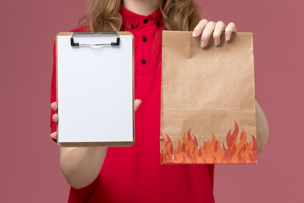 Front close view female courier in red uniform holding notepad and food package on light pink, job uniform service worker delivery