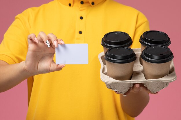 Front close view female courier in yellow uniform yellow cape holding coffee cups witj white card on pink background uniform delivery work color job