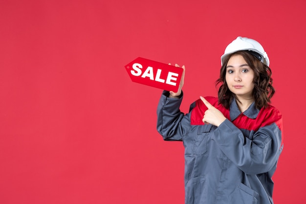 Front close view of female worker in uniform wearing hard hat and pointing sale icon on isolated red wall