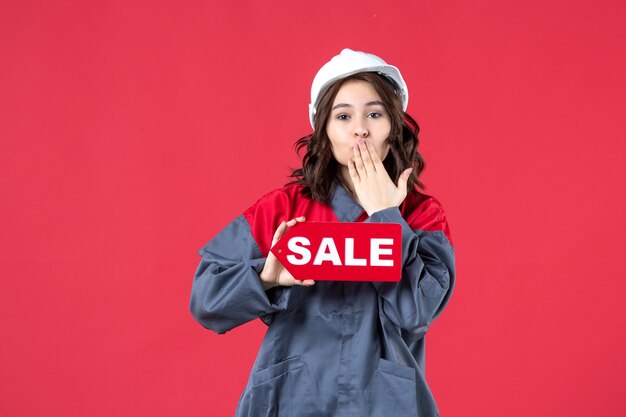 Front close view of female worker in uniform wearing hard hat showing sale icon and making kiss gesture on isolated red wall