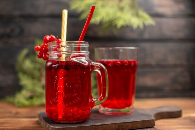 Front close view of fresh currant juice in a glass and a cup served with tube on a wooden cutting board on a brown table