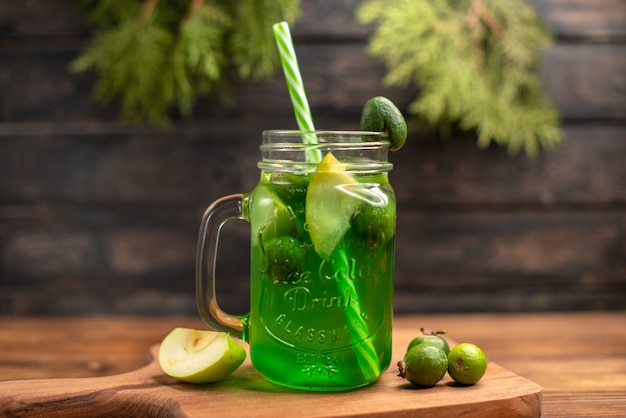 Front close view of fresh fruit juice in a glass served with tubes and apple limes feijoas on a wooden cutting board on a brown table