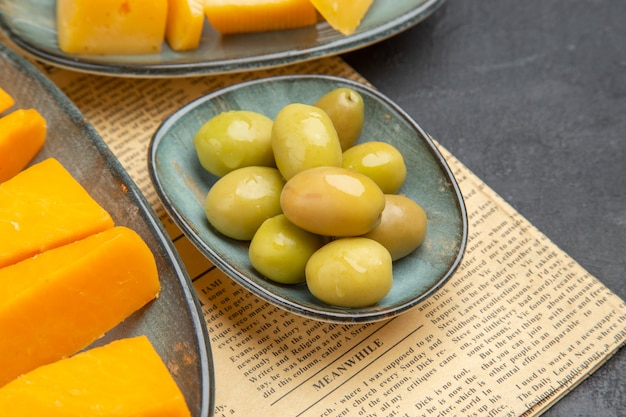 Front close view of fresh various sliced cheese and green olives on an old newspaper on a black background