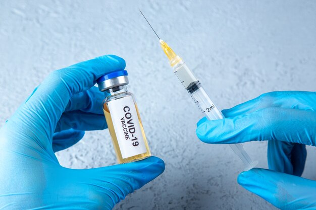 Front close view of glove wearing hand holding full syringe and covid- vaccine on gray sand background