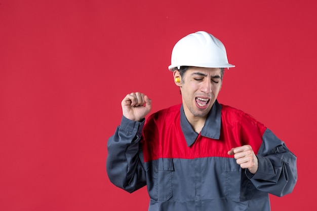 Vista frontale ravvicinata di un giovane lavoratore emotivo felice in uniforme con elmetto e che indossa tappi per le orecchie sul muro rosso isolato