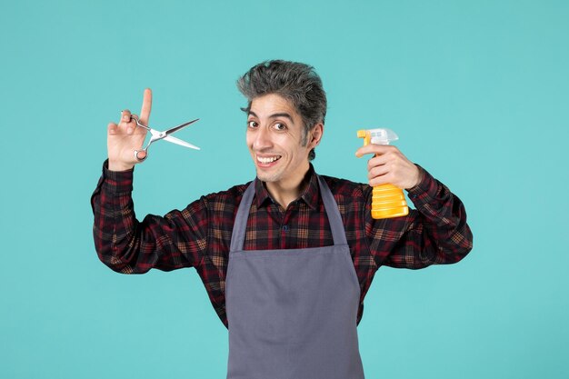 Front close view of happy excited male barber wearing gray apron and holding scissors spray bottle on soft blue color background