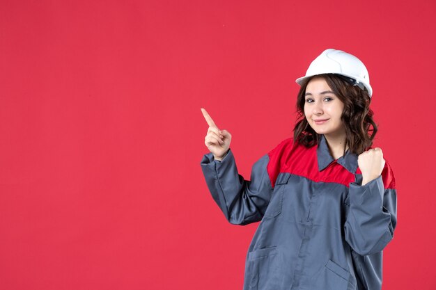 Front close view of happy smiling female builder in uniform with hard hat and pointing up on isolated red wall