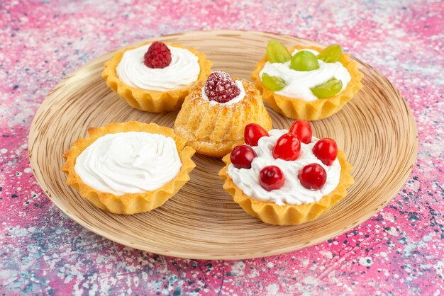 Front close view little cakes with fresh cream and fruits on the bright surface cookie 