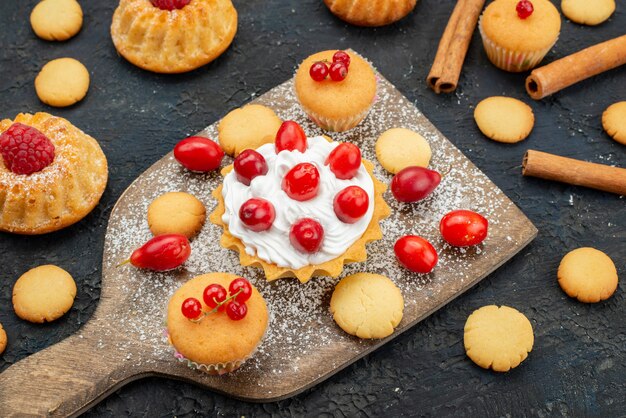 Front close view little yummy cakes with cream cinnamon and fresh fruits on the dark desk sweet biscuit cake dessert fruit berry