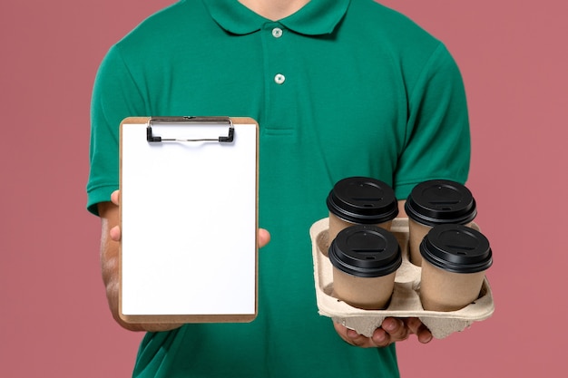 Front close view male courier in green uniform holding brown coffee cups and notepad on pink desk