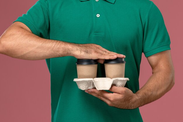 Front close view male courier in green uniform holding brown delivery coffee cups on the pink background 
