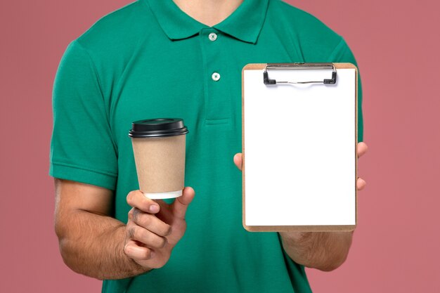 Крупным планом вид спереди мужчина-курьер в зеленой форме, держащий чашку кофе и блокнот на светло-розовом столе