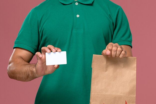 Front close view male courier in green uniform holding food package and card on pink background  