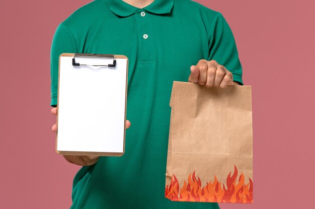 Front close view male courier in green uniform holding food package and notepad on light-pink background   