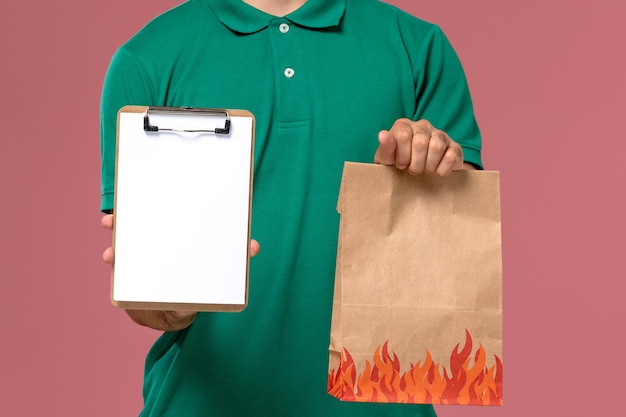 Front close view male courier in green uniform holding food package and notepad on light-pink background   