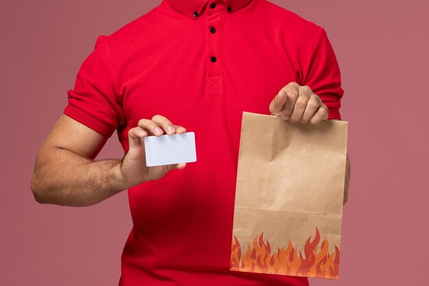 Front close view male courier in red uniform and cape holding food package and card on pink wall