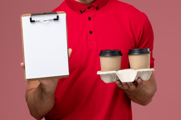 Front close view male courier in red uniform holding brown delivery coffee cups and notepad on light-pink desk worker