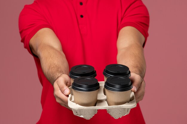 Front close view male courier in red uniform holding brown delivery coffee cups on pink wall 