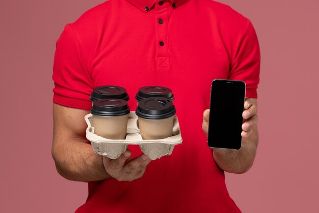 Front close view male courier in red uniform smiling and holding delivery coffee cups with phone on pink wall 