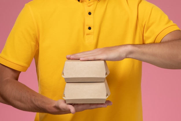 Front close view male courier in yellow uniform and cap with little delivery food packages on his hands on the pink wall