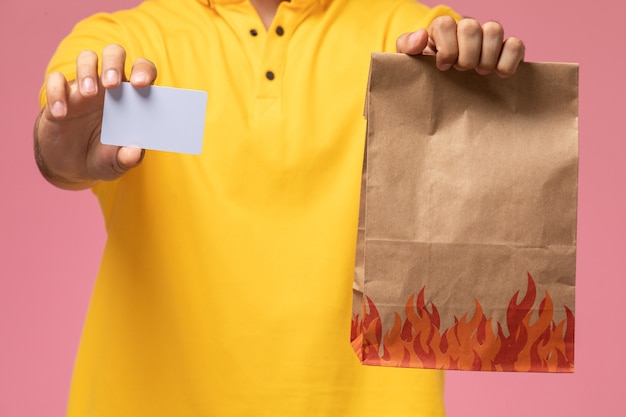 Front close view male courier in yellow uniform holding grey card and food package on the pink desk 