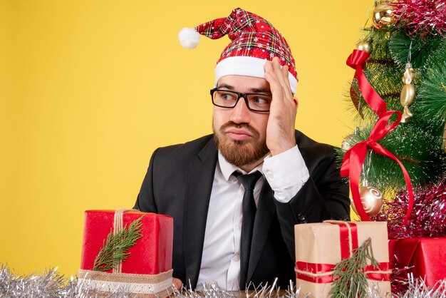 Front close view male worker sitting around presents and little tree on the yellow 