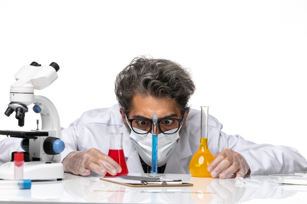 Front close view middle-aged scientist in special suit sitting with solutions and looking at them on a white background male virus science covid chemistry