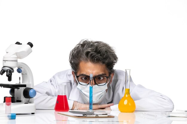 Вид спереди крупным планом ученый средних лет в специальном костюме, сидящий с растворами и смотрящий на них на белом столе, мужская вирусология, ковид-химия