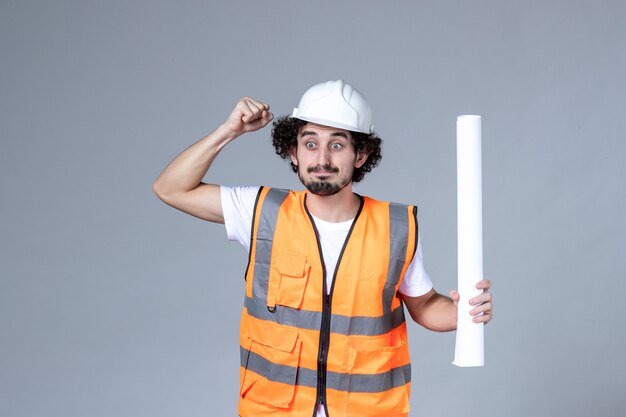 Front close view of nervous emotional male construction worker in warning vest with safety helmet and holding blank on gray wall