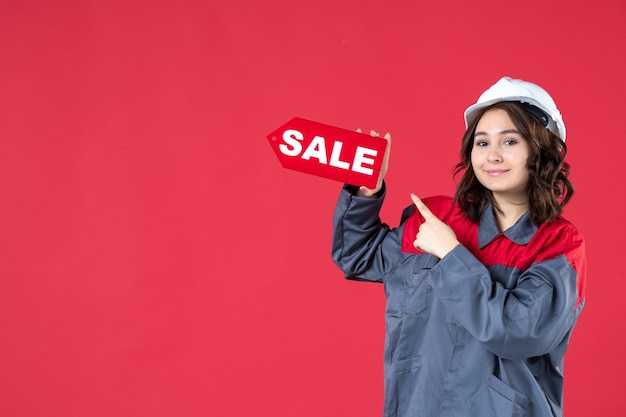 Бесплатное фото Крупным планом вид спереди улыбающейся работницы в униформе в каске и указывающей на значок продажи на изолированной красной стене