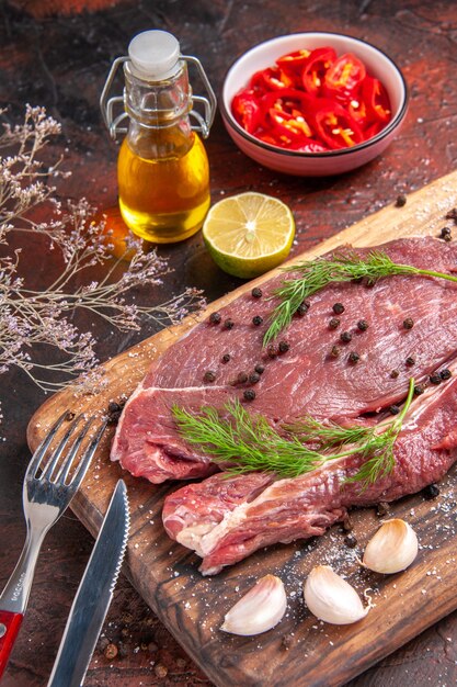 Front close view of red meat on wooden cutting board and garlic green fork and knife chopped pepper on dark background