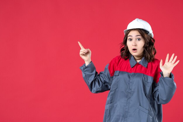 Front close view of shocked female builder in uniform with hard hat and pointing up on isolated red wall