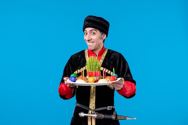 Front close view of smiling confident young guy in traditional dress holding tray filled with national confectionery on blue background