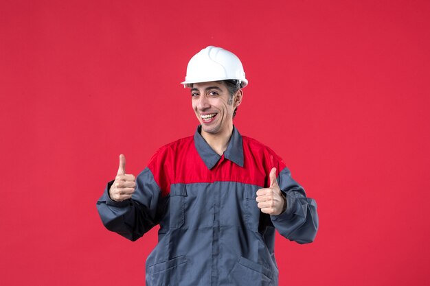 Front close view of smiling young builder in uniform wearing hard hat making ok gesture on isolated red wall