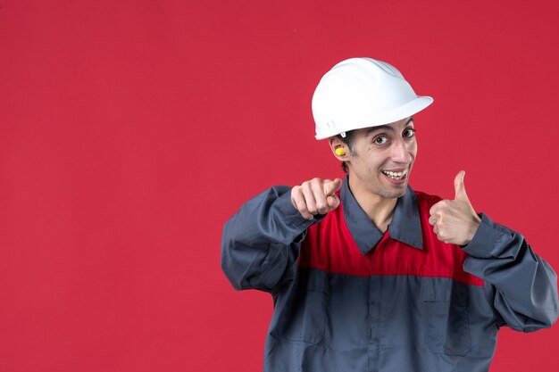 Front close view of smiling young worker in uniform with hard hat and wearing earplugs pointing forward making ok gesture on isolated red wall