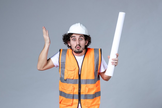 Front close view of worried construction worker in warning vest with safety helmet and holding blank on gray wall