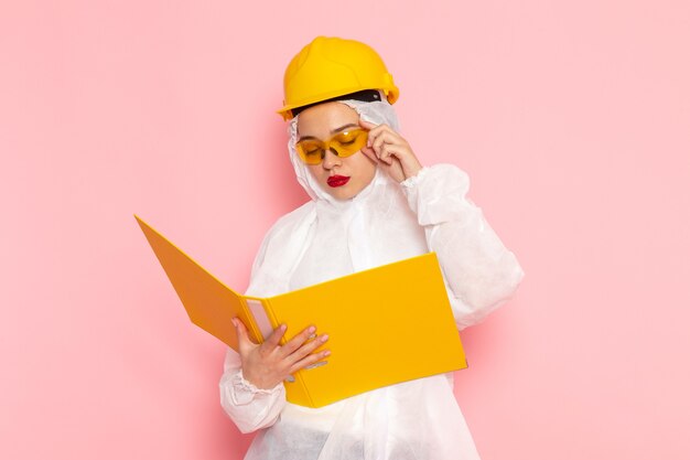 Front close view young beautiful female in special white suit wearing protective helmet reading a file on pink