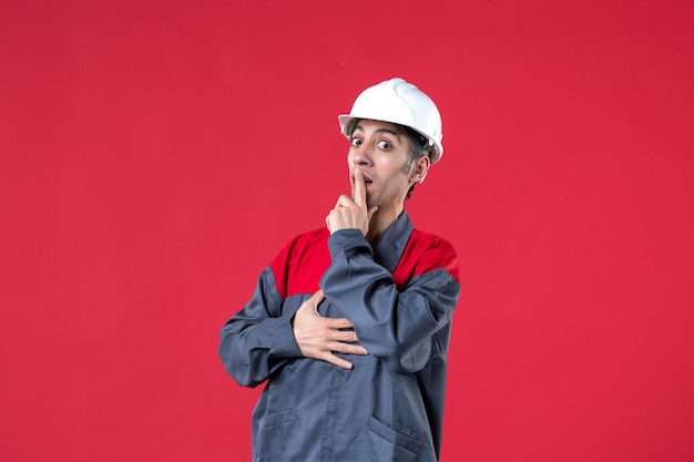 Front close view of young builder in uniform wearing hard hat feeling confused on isolated red wall