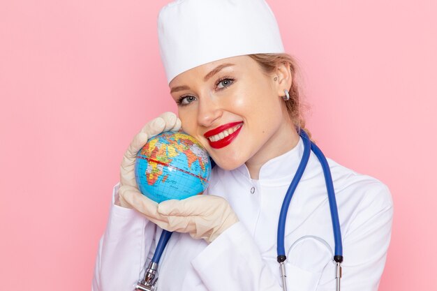 Вид спереди крупным планом молодая женщина-врач в белом медицинском костюме с синим стетоскопом, держащая глобус с улыбкой на розовом