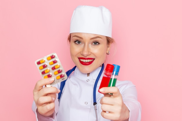 Front close view young female doctor in white medical suit with blue stethoscope holding pills and flasks on pink