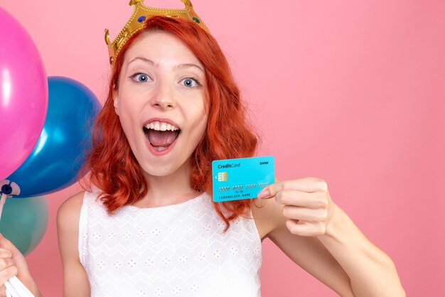 Front close view young female holding colorful balloons and bank card on pink 