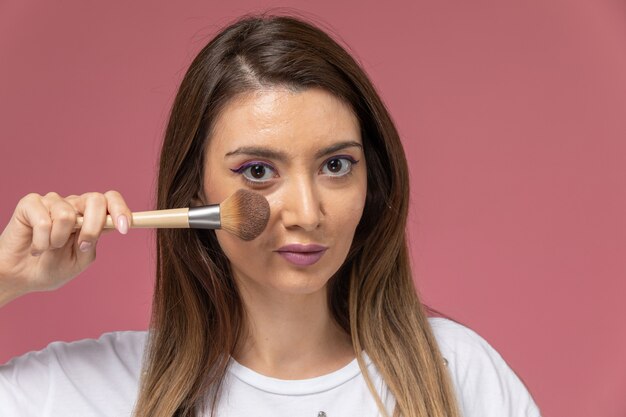 Front close view young female in white shirt holding make-up brush on pink, model woman pose beauty