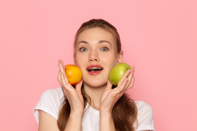 Front close view young female in white t-shirt holding fresh green apple with peach on light-pink wall