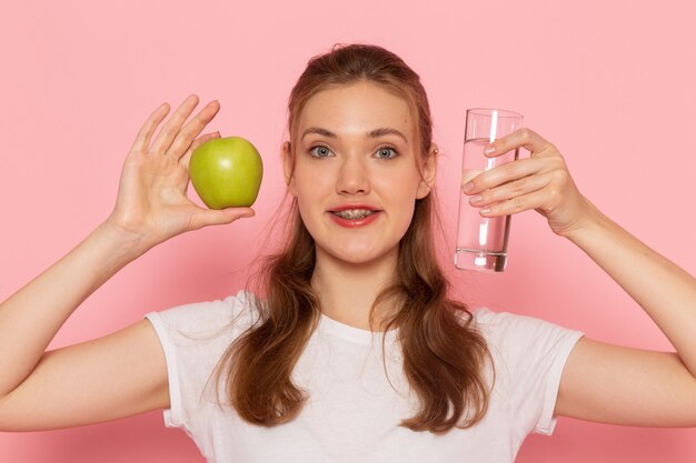 Front close view young female in white t-shirt holding green apple and glass of water on pink wall