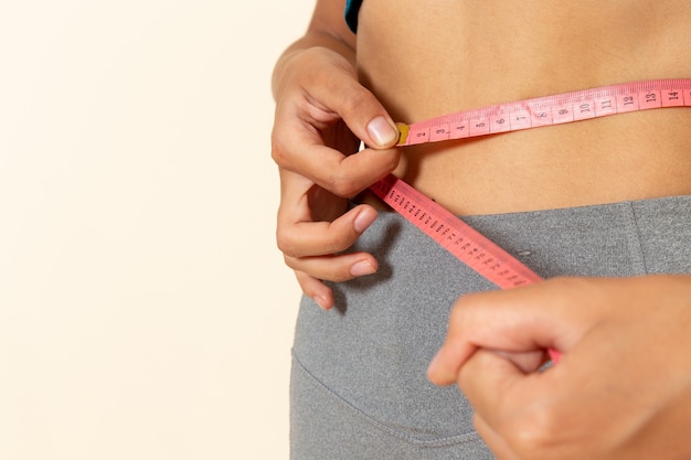 Front close view of young female with fit body in blue shirt measuring her waist on light white wall